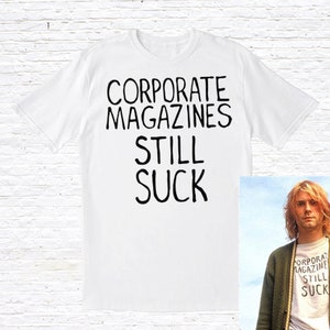 May include: White t-shirt with the black text "CORPORATE MAGAZINES STILL SUCK". A small image of a person wearing the same shirt is in the bottom right corner. The shirt has short sleeves and a crew neck.