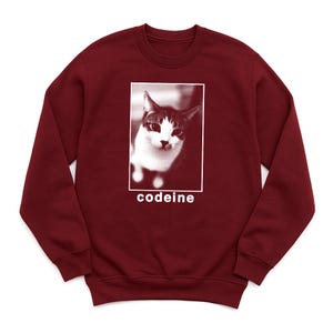 Puede incluir: Una sudadera burdeos con cuello redondo con una imagen en blanco y negro de un gato dentro de un rectángulo blanco. La palabra "codeine" está impresa en blanco debajo de la imagen. La sudadera está hecha de un material suave.