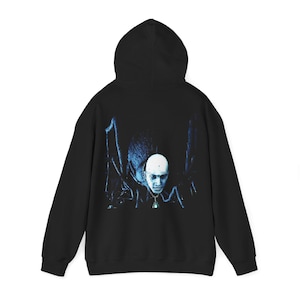Puede incluir: Sudadera con capucha negra con un gráfico azul y blanco de una cara pálida y un colgante. El gráfico tiene un fondo oscuro y sombreado. La sudadera tiene una capucha con cordón y mangas largas.