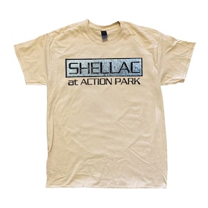 Könnte beinhalten: Ein beiges T-Shirt mit kurzen Ärmeln und einer rechteckigen Grafik auf der Brust. Die Grafik zeigt das Wort SHELLAC in einer abgenutzten schwarzen Schrift in einem Kasten, mit dem Text at ACTION PARK in schwarzer Schrift direkt darunter.
