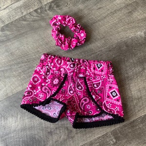 May include: A pink paisley shorts set with a matching scrunchie. The shorts have a black trim and a vibrant pink, white, and black paisley pattern. The scrunchie complements the shorts, offering a coordinated aesthetic. The set is designed for comfort and style.