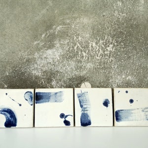 Op de afbeelding: Vijf witte keramische tegels met blauwe abstracte penseelstreken en spatten. De tegels zijn in een rij gerangschikt.