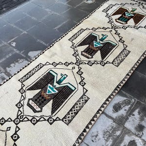 May include: A cream-coloured runner rug with a repeating geometric pattern featuring stylised eagle designs in black, brown, and teal. The rug has a fringed edge and is placed on a dark grey tiled surface.