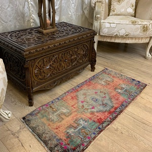 May include: A rectangular area rug featuring a faded, distressed pattern in shades of coral, sage green, and slate blue. The rug has a central geometric medallion, intricate border details, and short fringe along the edges, laid flat on a wooden floor.