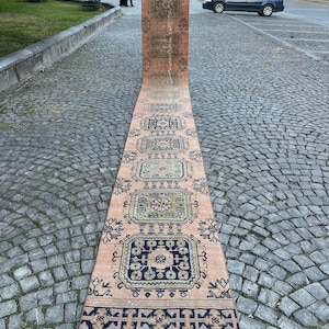 Puede incluir: Una alfombra larga y rectangular con un intrincado patrón geométrico. La alfombra presenta un fondo melocotón descolorido con detalles en azul marino, verde y crema. La alfombra se exhibe al aire libre sobre una superficie adoquinada.