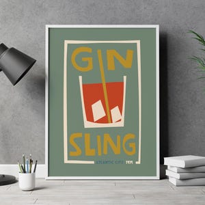 Puede incluir: Una l&aacute;mina enmarcada con fondo verde salvia que presenta una ilustraci&oacute;n estilizada de un c&oacute;ctel rojo en un vaso con cubitos de hielo y una pajita amarilla. El texto GIN SLING est&aacute; impreso en amarillo, con ATLANTIC CITY 1919 en la parte inferior.
