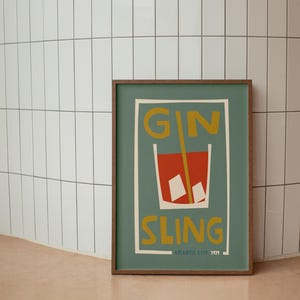 Puede incluir: Un p&oacute;ster rectangular enmarcado con fondo verde apagado que presenta una ilustraci&oacute;n estilizada de un c&oacute;ctel rojo en un vaso con cubitos de hielo y una pajita amarilla. El texto GIN SLING est&aacute; impreso en amarillo, con ATLANTIC CITY 1919 abajo.