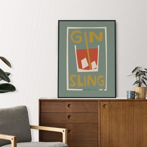 Puede incluir: Una l&aacute;mina de arte enmarcada con fondo verde salvia que presenta una ilustraci&oacute;n estilizada de un c&oacute;ctel rojo en un vaso con cubitos de hielo y una pajita amarilla. El texto GIN SLING est&aacute; impreso en amarillo, con ATLANTIC CITY 1974 debajo.