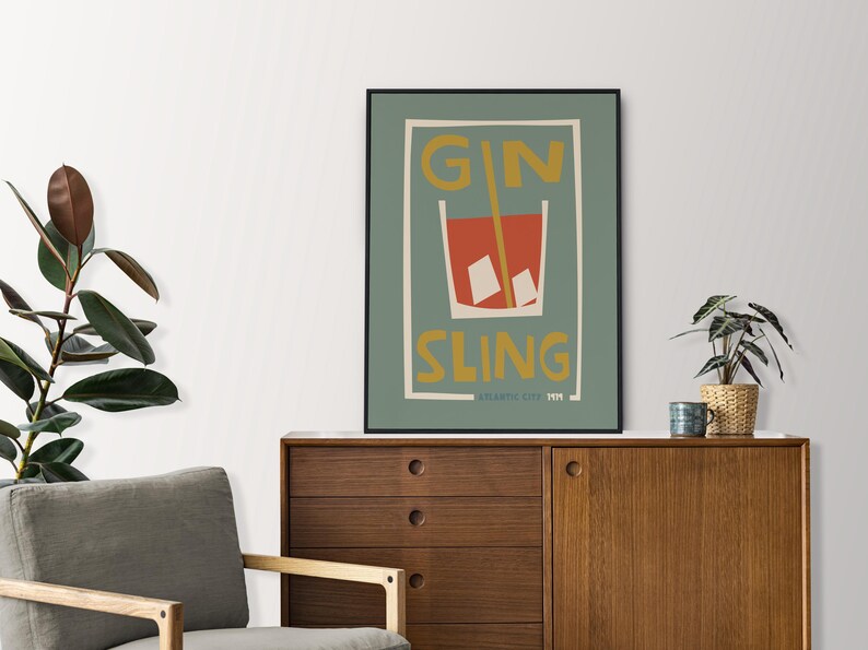 Puede incluir: Una l&aacute;mina de arte enmarcada con fondo verde salvia que presenta una ilustraci&oacute;n estilizada de un c&oacute;ctel rojo en un vaso con cubitos de hielo y una pajita amarilla. El texto GIN SLING est&aacute; impreso en amarillo, con ATLANTIC CITY 1974 debajo.