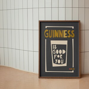 Op de afbeelding: Een ingelijste poster met een zwarte achtergrond en witte tekst die "GUINNESS IS GOOD FOR YOU DUBLIN" luidt. De tekst is in een vetgedrukt, gestileerd lettertype. Een witte omtrek van een glas Guinness bevindt zich in het midden van de poster.