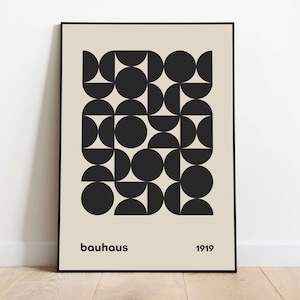 Op de afbeelding: Een rechthoekige wandkunstprint in een zwarte lijst met een beige achtergrond en een centraal raster van zwarte geometrische vormen, waaronder cirkels en halve cirkels. De tekst "bauhaus" staat linksonder en "1919" rechtsonder.