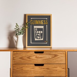 Op de afbeelding: Een ingelijst print met een zwarte achtergrond en gouden tekst die "GUINNESS IS GOOD FOR YOU" zegt met een glas Guinness eronder. De print staat op een houten plank met een witte kan en een groene plant.