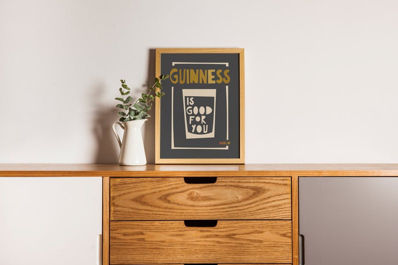 Op de afbeelding: Een ingelijst print met een zwarte achtergrond en gouden tekst die "GUINNESS IS GOOD FOR YOU" zegt met een glas Guinness eronder. De print staat op een houten plank met een witte kan en een groene plant.