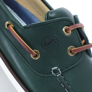 Puede incluir: Primer plano de un zapato n&aacute;utico de cuero verde oscuro con cordones marrones y ojales de metal dorado. El zapato tiene una suela blanca y un peque&ntilde;o logotipo de ancla en el lateral.
