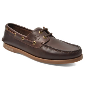 May include: A brown leather boat shoe with a light brown sole. The shoe has a lace-up design with gold-coloured eyelets and the brand name "SEAJURE" on the side. The shoe has a classic, casual style.