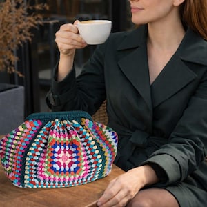 Può includere: Una pochette all'uncinetto fatta a mano con un vivace motivo geometrico. La borsa presenta un mix di colori vivaci tra cui rosa, blu, verde e giallo. La borsa è appoggiata su un tavolo di legno.