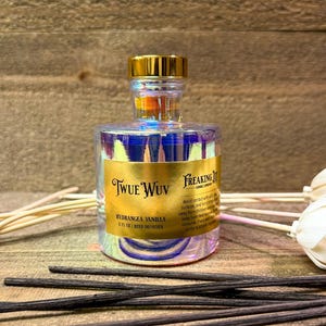 May include: A clear glass reed diffuser bottle with a gold cap and a label that reads "Twue Wuv Hydrangea Vanilla". The bottle is surrounded by black reed sticks and other decorative elements. The diffuser is 5 fl oz.