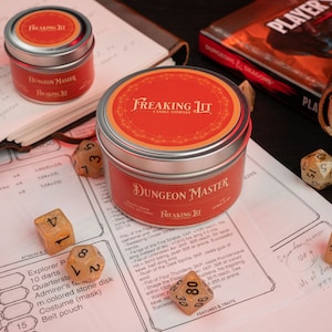 May include: Two silver candle tins with red labels that say "Freaking Lit Candle Company" and "Dungeon Master" on a tabletop with dice and a Dungeons and Dragons rulebook.