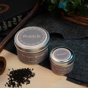 May include: Two silver tins of The Grey Wizard candle by Freaking JIT, with a dark wooden pipe and a copy of The Fellowship of the Ring. The tins have a gray label with ornate detailing. Loose black tea leaves are scattered nearby.