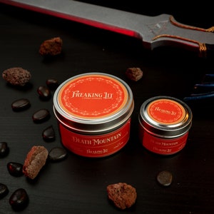 May include: Two candles in silver tins with red labels. The larger candle is labeled "Death Mountain" and the smaller candle is labeled "Death Mountain". Both labels have the text "Freaking Lit Candle Co."