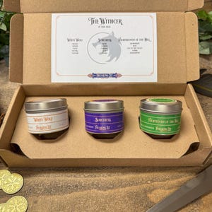 May include: A brown cardboard box with three small metal tins inside. The tins are labeled "White Wolf", "Sorceress", and "The Witcher of the Hill". The box is open and the tins are visible. The box is sitting on a wooden surface.