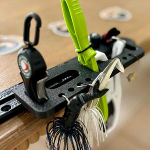 May include: A black fishing lure organizer with a variety of fishing lures. The organizer has a bright green tool and a black clip. The lures include a black and blue jig, and white and silver lures.