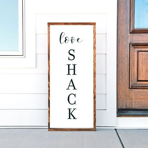 Pode incluir: Uma placa de madeira com as palavras "love shack" escritas em letras cursivas e maiúsculas pretas. A placa é marrom e tem um fundo branco.