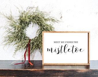May include: A wooden framed sign with the text "Meet me under the mistletoe" in black script. A green wreath with red accents and a red ribbon hangs on the wall behind the sign. The sign and wreath are on a dark wooden surface.
