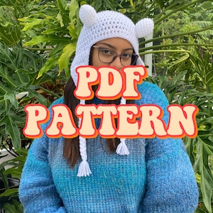 May include: A white crocheted hat with ear flaps and a white pom-pom on each ear flap. The hat is being worn by a person with brown hair and glasses. The text "PDF PATTERN" is superimposed on the image.
