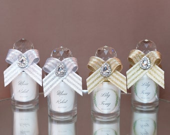 May include: Four small glass candle holders with crystal toppers, adorned with ribbons and jewel embellishments. Two have silver ribbons, and two have gold ribbons. Each candle has a label with names and a date. The candles are on a reflective surface.