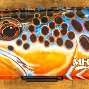 May include: Rectangular fly fishing box with a colorful design of a brown trout's head. The box features orange, blue, and black patterns, with the brand name "MFC" and a fish logo on the side. The box has a black latch.