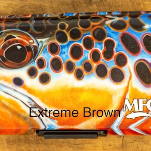 May include: A rectangular fly fishing box featuring a detailed graphic of a brown trout eye and scales in vibrant orange, blue, and black. The text Extreme Brown and the MFC logo with a fish icon are printed on the lower right side of the case.