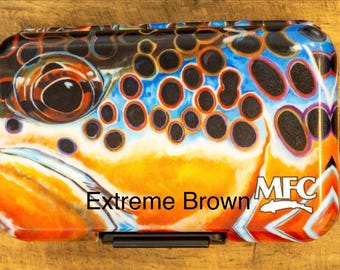 May include: A rectangular fly fishing box featuring a detailed graphic of a brown trout eye and scales in vibrant orange, blue, and black. The text Extreme Brown and the MFC logo with a fish icon are printed on the lower right side of the case.