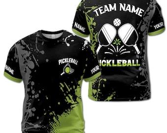 May include: Black and green pickleball t-shirt with a splatter paint design. The shirt has the text "TEAM NAME" on the back and "PICKLEBALL" on the front. The shirt also has a graphic of two pickleball paddles and a ball.