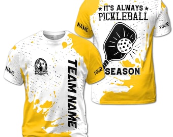 May include: Yellow and white t-shirt with a black and white pickleball paddle graphic. The text "It's Always Pickleball" and "Your Season" are on the shirt. The text "Team Name" is on the side of the shirt.