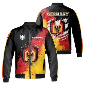 Könnte beinhalten: Schwarze Bomberjacke mit einem farbenfrohen Design der deutschen Flagge. Die Jacke hat einen Reißverschluss und zwei Taschen. Die Rückseite der Jacke zeigt das Wort "DEUTSCHLAND" und einen stilisierten deutschen Adler.