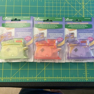 May include: Three packages of Clover Desk Needle Threaders. Each package is a different color: green, orange, and purple. The packages feature the Clover logo and the text "Desk Needle Threader" in white. The text "Thread your needles quickly with the help of Clover's Desk Needle Threader!" is also included on each package.