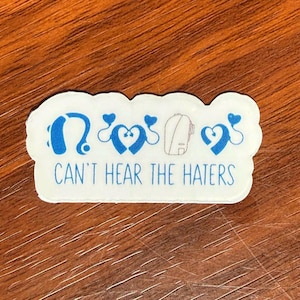 May include: A white sticker with a blue design and text. The design includes hearing aids and hearts. The text reads "CAN'T HEAR THE HATERS" in blue. The sticker is on a wooden surface.