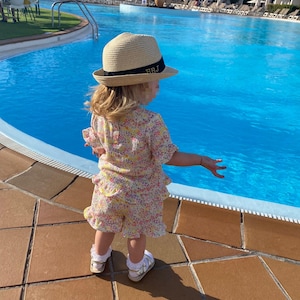Può includere: Un bambino piccolo con un cappello di paglia con le iniziali "BRJ" ricamate sulla fascia, un vestito a stampa floreale e sandali bianchi è in piedi vicino a una piscina.