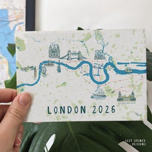 Puede incluir: Una postal rectangular con un mapa de Londres con ilustraciones de lugares emblemáticos como el London Eye y el Tower Bridge. El texto "LONDON 2026" está impreso en verde. La postal se sostiene frente a una hoja verde.