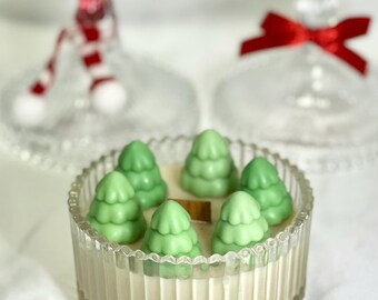 Puede incluir: Un tarro de vela de vidrio transparente con un diseño acanalado, lleno de una vela blanca y cubierto con decoraciones de cera en forma de árbol de Navidad verde.