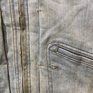 May include: A light blue denim jacket featuring a central vertical zipper closure with brown stitching, vertical seams, and a single chest pocket on the right side. The fabric shows a light wash finish with a small hole near the pocket seam.