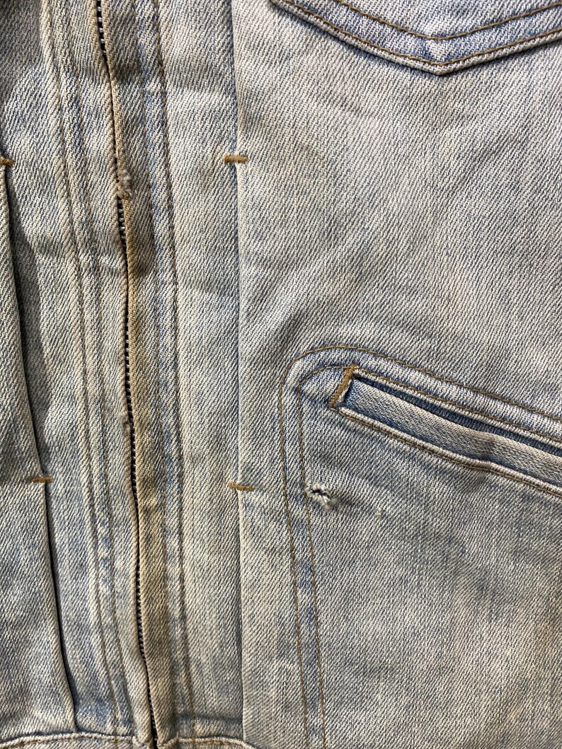 May include: A light blue denim jacket featuring a central vertical zipper closure with brown stitching, vertical seams, and a single chest pocket on the right side. The fabric shows a light wash finish with a small hole near the pocket seam.