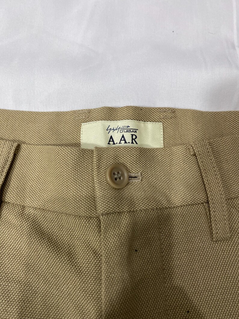Puede incluir: Primer plano de un pantal&oacute;n beige con cierre de bot&oacute;n y una etiqueta que dice "A.A.R. Urban". La tela tiene un tejido texturizado. El pantal&oacute;n probablemente forma parte de una colecci&oacute;n de ropa.