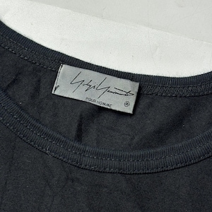 May include: Close-up of a black t-shirt with a crew neck. The label features the brand name "Yohji Yamamoto POUR HOMME" in a stylized font. The shirt is made of a soft, dark fabric.