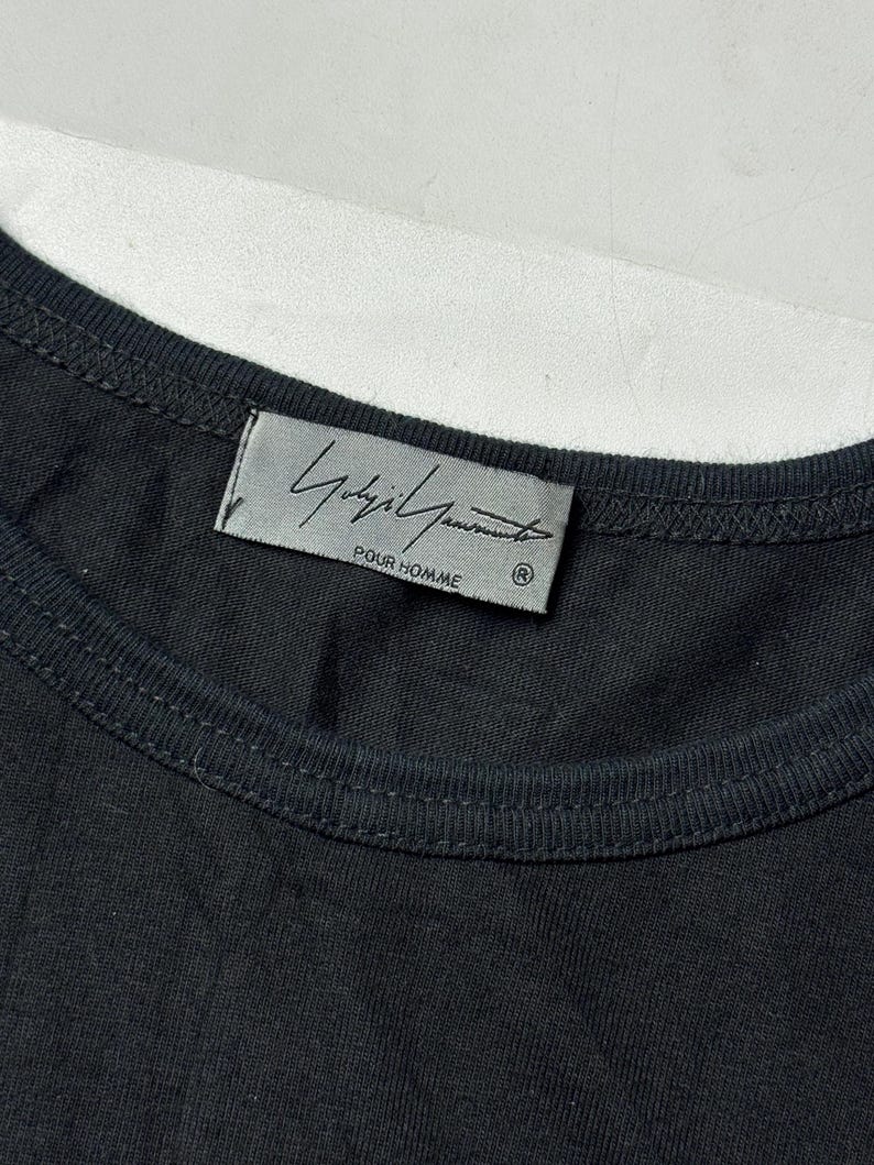 May include: Close-up of a black t-shirt with a crew neck. The label features the brand name "Yohji Yamamoto POUR HOMME" in a stylized font. The shirt is made of a soft, dark fabric.