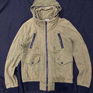 May include: Olive green hooded jacket with a full-length zipper, two chest pockets, and two lower pockets. The jacket has a black ribbed hem and cuffs. The jacket has a hood and a high collar. The jacket is made of a soft, textured material.