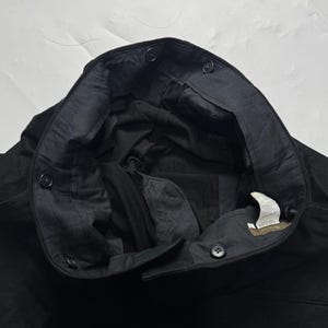 Puede incluir: Primer plano de pantalones negros con cierre de bot&oacute;n y forro interior oscuro. Los pantalones tienen varios botones a lo largo de la cintura y una etiqueta en el interior. La tela parece ser un material resistente.