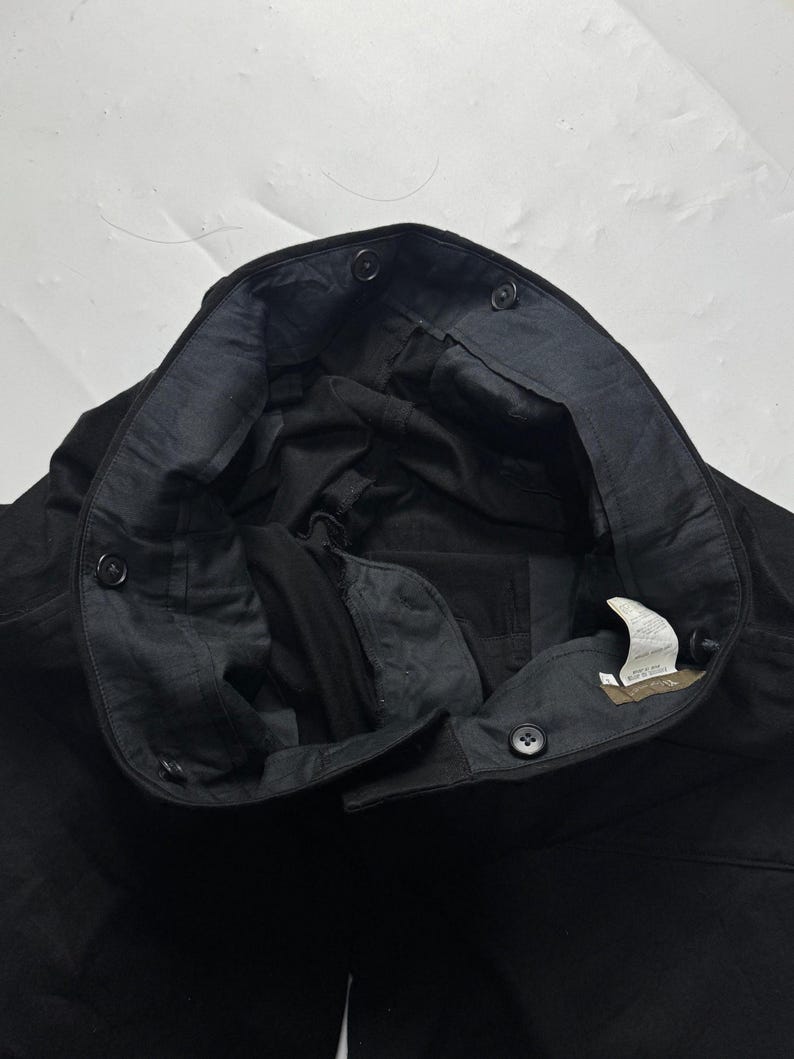 Puede incluir: Primer plano de pantalones negros con cierre de bot&oacute;n y forro interior oscuro. Los pantalones tienen varios botones a lo largo de la cintura y una etiqueta en el interior. La tela parece ser un material resistente.