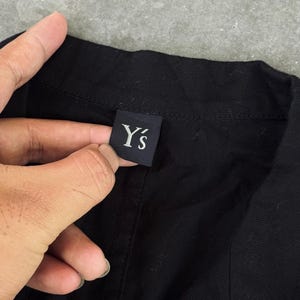 May include: Close-up of a black garment with a label that reads "Y's" in white lettering. The label is rectangular and appears to be made of a dark fabric. The garment is a solid black color.